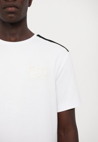 Biała koszulka z krótkim rękawem z okrągłym dekoltem, z czarnym akcentem na ramieniu i złotym logo EA7 Emporio Armani. Gładka faktura.
