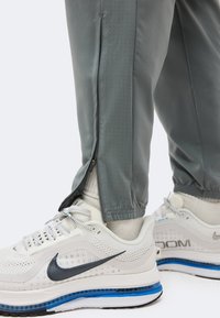 Γκρίζο παντελόνι με μερικώς ανοιχτό πλαϊνό φερμουάρ πάνω από λευκά αθλητικά Nike Air Zoom με μπλε σόλα, φορεμένα με λευκές κάλτσες.