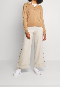 Beige gebreide polo met lange mouwen, met een kraag en knoopsluiting, gecombineerd met brede crèmekleurige broek met knoopaccenten aan de zijkanten.
