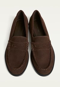 Braune Wildleder-Slipper mit einem Slipper-Design, das kontrastierende Nähte und eine abgerundete Spitze aufweist, auf einer schwarzen, strukturierten Sohle.