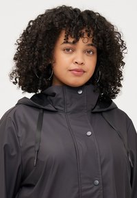 A young woman with curly hair wears a black hooded jacket, featuring a zip and snap closure, looking directly at the viewer with a neutral expression.