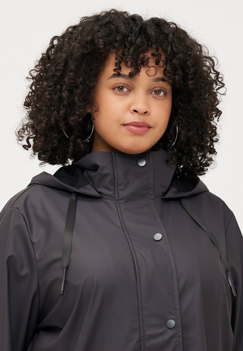 Een jonge vrouw met krullend haar draagt een zwarte gewatteerde jas met capuchon, voorzien van een ritssluiting en drukknopen, en kijkt met een neutrale uitdrukking recht in de camera.