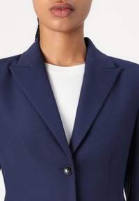 Marineblazer mit Reverskragen, einzelnem schwarzen Knopfverschluss und glatter Textur, kombiniert mit einem weißen Rundhals-T-Shirt darunter.