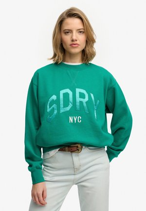 Zielona bluza z dużym nadrukiem "SDRY NYC" wykonanym z teksturowanego, odblaskowego materiału. Posiada okrągły dekolt i długie rękawy, zestawiona z jasnymi dżinsami.
