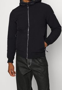 Sort lynlåsjakke med hætte, der har en glat tekstur, forlommer og elastiske manchetter. Kombineret med mørke bukser med kontrastsyninger.