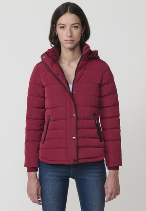 Chaqueta acolchada burdeos con capucha, textura acolchada, bolsillos con cremallera y detalles en cremallera negra. Corte ligeramente corto, combinada con jeans azules.