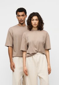 Δύο μοντέλα φορούν υπερμεγέθη, μπεζ T-shirts με κοντά μανίκια, συνδυασμένα με ανοιχτόχρωμο μπεζ παντελόνι φόρμας, με ελαστική μέση και άνετη εφαρμογή.