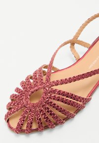 Offene Sandalen aus geflochtenem, roségoldenen Material mit einer braunen Sohle. Sie verfügen über sich kreuzende Riemen und einen gemusterten Akzent an der Vorderseite.