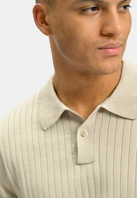 Beige ribbad polo skjorta med en mjuk textur, tre-knappars stängning och en klassisk krage, som framhäver subtila vertikala ränder.