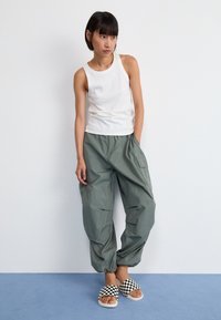 White ribbed tank top, loose dark green cargo pants with pockets, and black-and-white checkered slide sandals on a light blue floor.