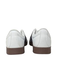 Vita lädersneakers med bruna gummisålar, med en slät yta och två svarta ränder på varje sida, sett bakifrån.