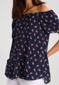 Off-shoulder mörkblå blus i lätt tyg, med ett flamingotryck i rosa och grönt, och korta, svallande ärmar.