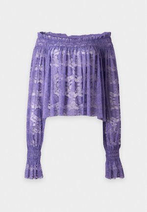 Frau trägt eine transparente lila schulterfreie Spitzenbluse mit langen Ärmeln, schwarze knielange Shorts und schwarze Sandalen mit Absatz.