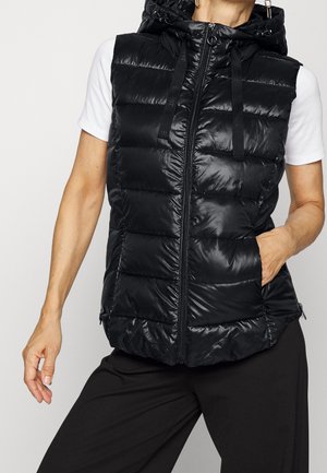 Femme portant un gilet matelassé noir sans manches à capuche sur une chemise blanche à manches courtes, une main dans la poche du gilet.
