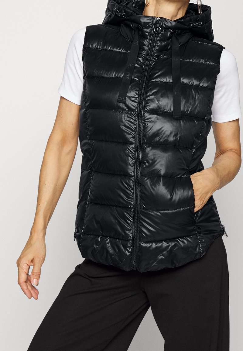 Femme portant un gilet matelassé noir sans manches à capuche sur une chemise blanche à manches courtes, une main dans la poche du gilet.
