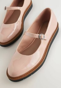 Chaussures Mary Jane beige brillantes avec une boucle argentée, une épaisse bordure marron et des semelles en caoutchouc noires sur une surface blanche.