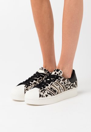 Personne portant des baskets imprimé léopard noir et blanc avec des lacets noirs et des semelles blanches sur fond blanc.