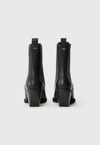 Par svart skinn ankelstøvler med blokkhæler, ribbede elastiske sidepaneler og pull tabs, vist fra baksiden på hvit bakgrunn.