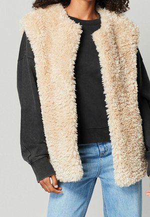 Personne portant un gilet beige duveteux sans manches par-dessus une chemise noire à manches longues et un jean bleu clair sur un fond uni.