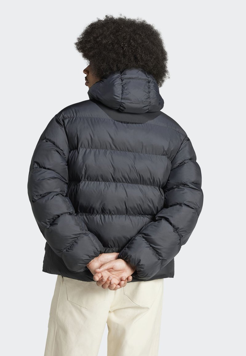 Schwarze, puffy Jacke mit hohem Kragen, gesteppter Optik und elastischen Manschetten, getragen über hellfarbigen Hosen, die eine strukturierte Oberfläche zeigen.