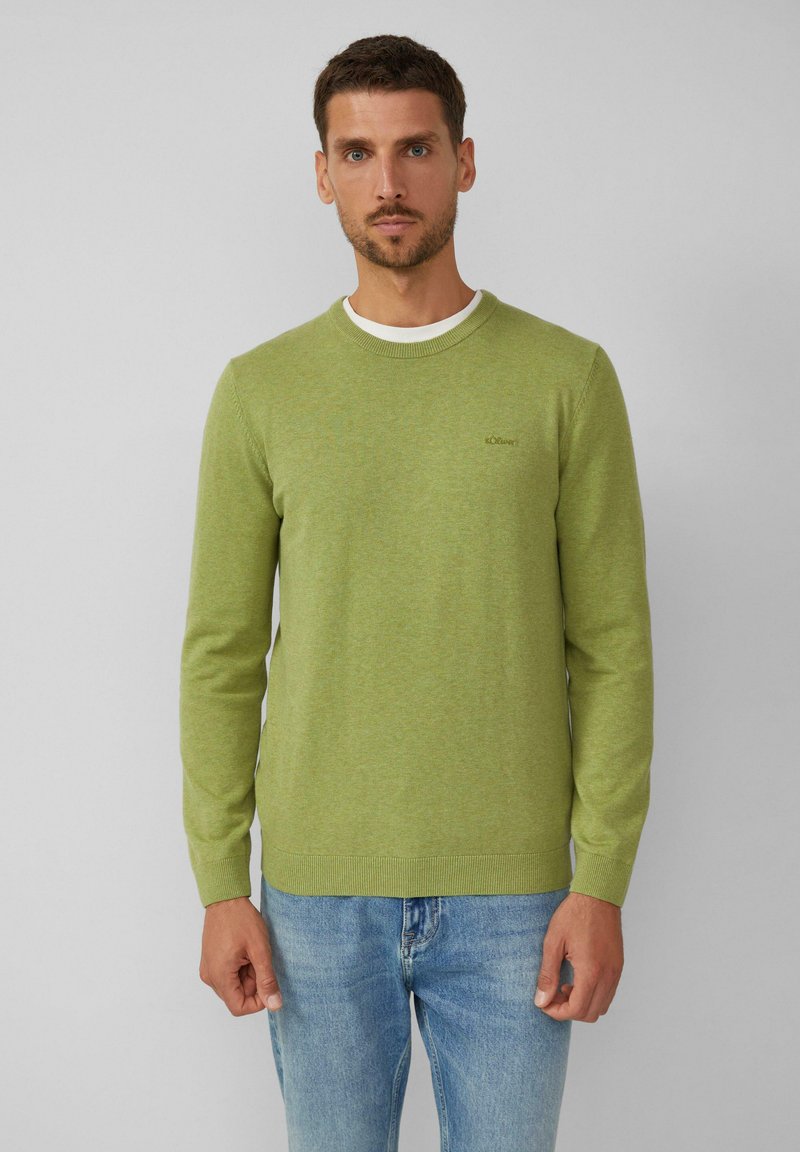 Hellgrüner Strickpullover mit Rundhalsausschnitt und gerippten Bündchen, versehen mit einem dezenten Logo-Detail. Kombiniert mit blauen Denim-Jeans.
