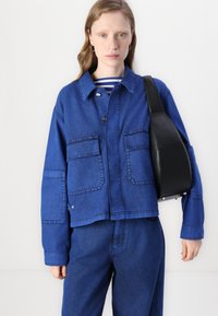 Blå denimjakke med store frontlommer, knappeåpning og en litt cropped passform, parret med en svart veske. Stripete skjorte synlig under.