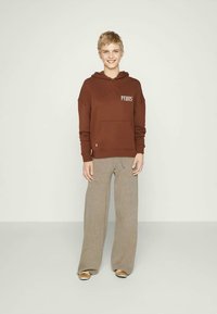 Brązowy kaptur z kieszenią z przodu i białym napisem, w połączeniu z jasnobeżowymi, szerokimi spodniami. Gładki materiał i luźny krój.