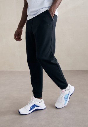 Person trägt schwarze Nike Jogginghosen und weiße Nike Sneakers mit blauen Akzenten, steht auf beigem Boden vor einer neutralen Wand.