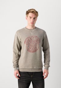 Grå hettegenser med et rundt rødt logo med teksten "TWENTY TWO SCOTCH & SODA AMSTERDAM". Lange ermer og ribbestrikkede mansjetter. Myk tekstur.