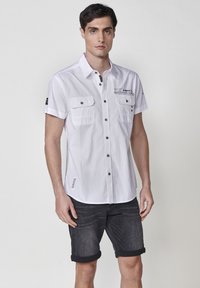 Camisa blanca de manga corta con dos bolsillos en el pecho, cierre de botones y detalles de costura negra. Se lleva con pantalones cortos de mezclilla oscura, con los puños ligeramente doblados.