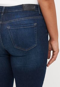 Dark blue denim jeans with a black leather patch at the waistband, featuring two back pockets with contrast stitching. Smooth texture.