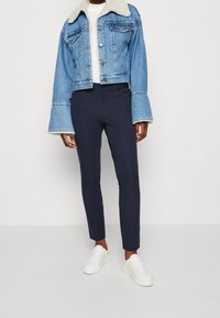 Person som bär en kort blå jeansjacka med vit fårskinnskrage och manschetter, marinblå slim-fit byxor, vita sneakers och vit topp.