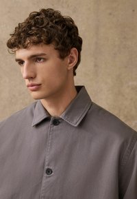 Szary płaszcz zapinany na guziki z usztywnionym kołnierzem, wykonany z gładkiego, matowego materiału. Dwa czarne guziki widoczne z przodu do zapięcia.