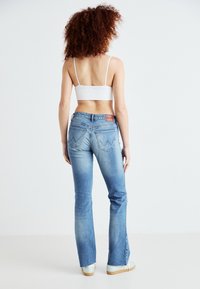 Jeans de mezclilla elástica azul con piernas acampanadas, cintura de altura media, detalles desgastados y bolsillos traseros con un distintivo diseño de costura en forma de V.