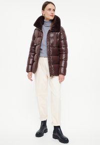 Brązowa kurtka puchowa z pluszowym kołnierzem, błyszczącym wykończeniem i zapięciem na zamek. Połączona z jasnymi spodniami i czarnymi botkami za kostkę.