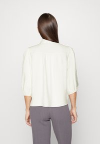White, lightweight blouse with a collar, three-quarter sleeves, and pleated back detail, paired with gray ribbed pants.