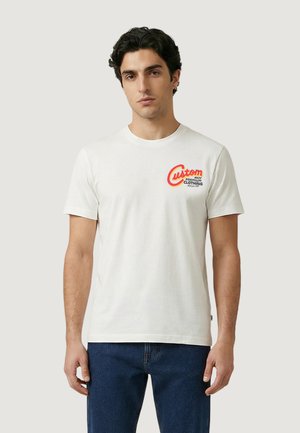 Giovane uomo in piedi che indossa una maglietta bianca con un logo rosso e giallo "Abbigliamento premium su misura" sul lato sinistro del petto e jeans blu.