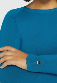 Teal randig långärmad topp med rund halsringning och knappdetalj vid manschetten. Mjuk textur, figurnära passform och synliga sömsömmar.