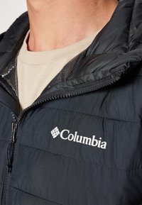Zwarte geïsoleerde jas met een ritssluiting aan de voorkant, met horizontale naden en het Columbia-logo in het wit. Mesh-accenten zichtbaar rond de kraag.