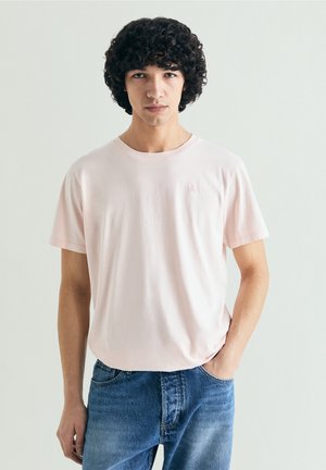 Joven con cabello oscuro y rizado, vestido con una camiseta rosa claro y jeans azules, posando frente a un fondo claro y liso.