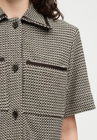 Femme portant une chemise à manches courtes à motif pied-de-poule noir et blanc avec des boutons noirs et une bordure foncée sur le col et les poches poitrine.