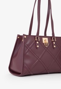 Bolso tote de cuero burdeos con herrajes dorados, patrón acolchado y asas dobles. Presenta un logo metálico decorativo en la parte frontal.