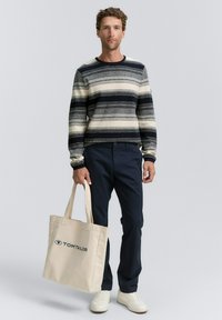 Man in een gestreepte marineblauwe en crèmekleurige trui, donkere broek en witte sneakers die een beige Tom Tailor tote bag vasthoudt op een effen achtergrond.
