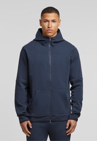 Mornarska zip-up dukserica izrađena od glatkog materijala, s visokim ovratnikom, velikim prednjim džepovima i opuštenim krojem s dugim rukavima.