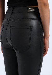 Zwarte nepleer jeans met een hoge taille, voorzien van twee achterzakken, subtiele stiksels en een merklabel op de taille.