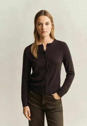 Vrouw met lang blond haar die een donkerkleurige vest met knopen en donkere broek draagt, staand met één hand in de zak tegen een effen achtergrond.