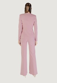 Donna in piedi di spalle, che indossa un blazer rosa su misura e pantaloni a gamba larga coordinati, con i capelli raccolti a metà in una coda di cavallo.