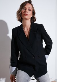 Černý dvouřadý blazer s klopami, s kontrastními šedými manžetami. Hladká textura a dobře padnoucí střih.