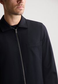 Chaqueta negra texturizada con cremallera frontal, cuello alzado y un bolsillo en el pecho. Fabricada con material transpirable, diseñada para un ajuste moderno.