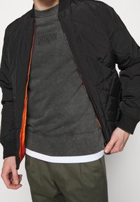 Czarna pikowana kurtka bomberka z pomarańczową podszewką, noszona na ciemnoszarej bluzie. U dołu bluzy widoczny biały brzeg.